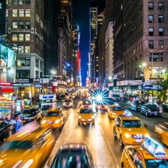 A busy city street with cars moving so fast they are blurred but the street has so many cars and it looks like New York so there's no way traffic isn't at like a complete standstill so that effect is probably misleading. Also, lots of taxis on the road.