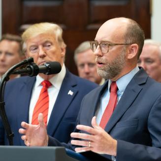 Russell Vought speaks into a microphone, with Donald Trump in the background.