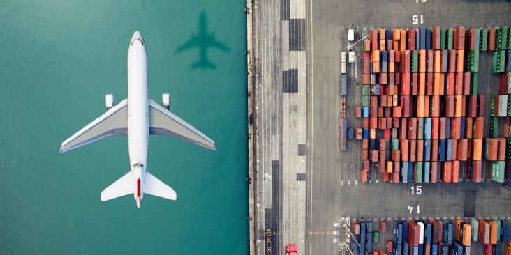 A plane flying over a dock, half of the ground is water and half is shipping containers