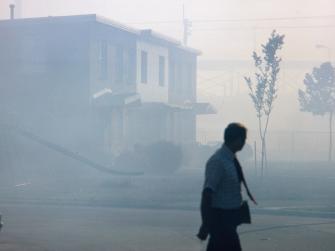 Smog clouds a city street, shrouding residential buildings and small trees as a person in a tie walks by.