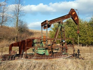 A weathered oil pumpjack with visible rust and peeling paint stands in a dry grassy field. Bare deciduous trees and evergreen shrubs surround the site, with rolling farmland and a partly cloudy blue sky in the background. The aging equipment suggests a low-producing or inactive oil well in a rural setting.